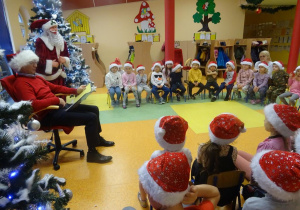 Grupa dzieci siedzi na krzesłach wokół pana Marka Cieślaka, który czyta dzieciom książkę. W tle dekoracja świąteczna.